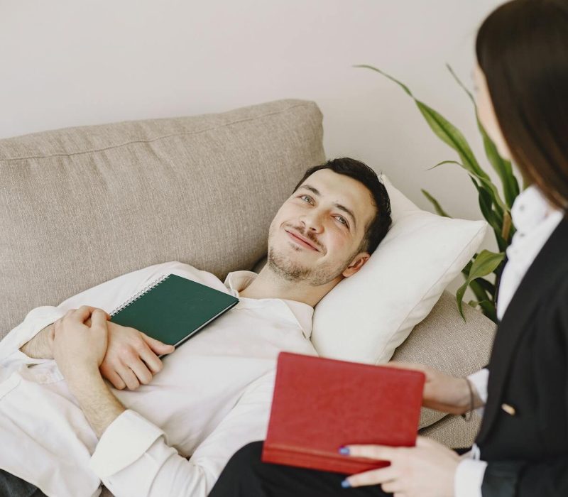 Positive man in formal wear lying on sofa with notebook in hands while smiling and talking with therapist in black suit