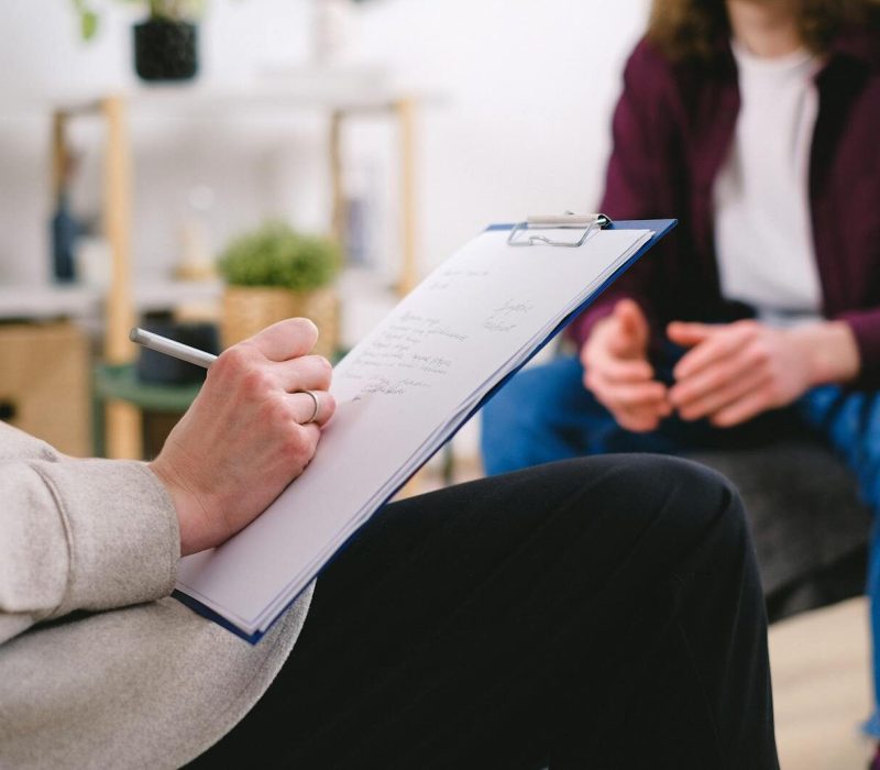 Close-up of a therapist writing notes on a clipboard while conversing with a patient.